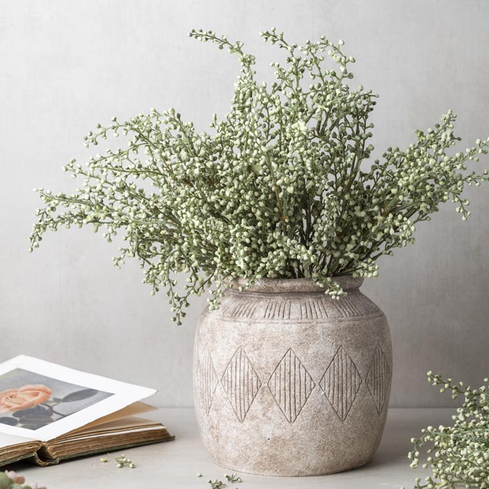 A artificial sage-colored heather stem arranged in a beige ceramic vase, with a soft focus background.