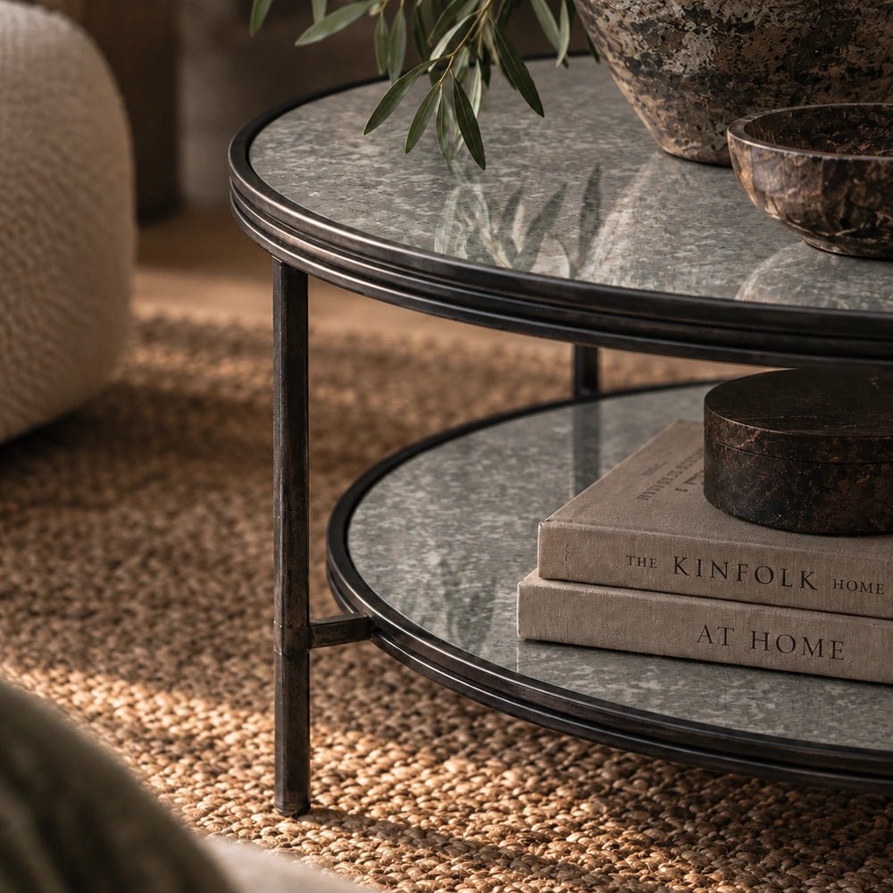 Close-up of the Leiden Metal Coffee Table showing its slim dark round frame, reflective two-tier surfaces, stacked books and softly styled living room.
