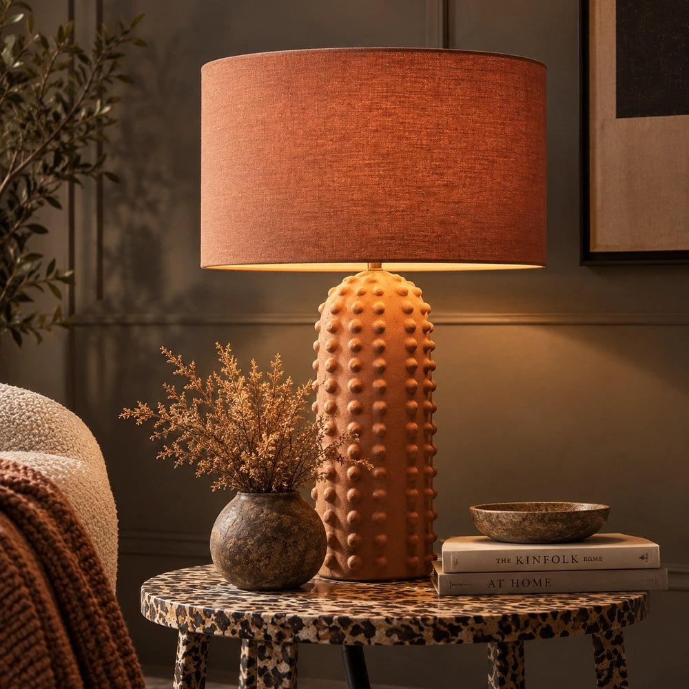 Maynard Terracotta Table Lamp with tall textured bobble base and wide fabric shade, illuminated on a patterned side table beside dried foliage and books.