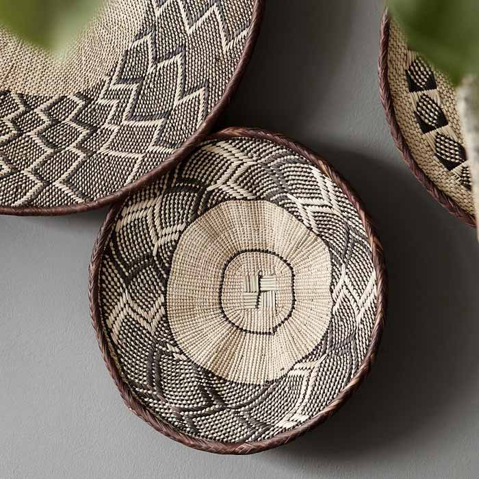 The Melina Basket, a circular shallow woven basket with a geometric tribal pattern, pictured in a group of three on a grey wall.
