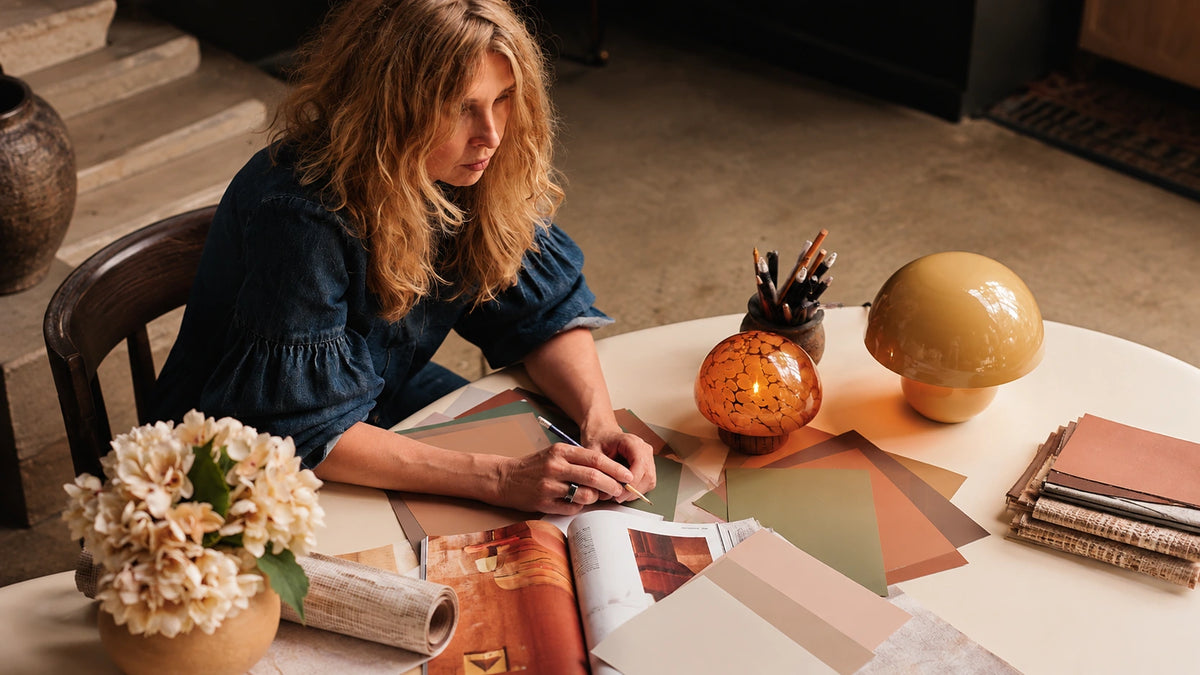 Abigail Ahern selecting warm neutral colour samples at a studio table with fabric swatches, an interiors book, flowers and sculptural table lamps.