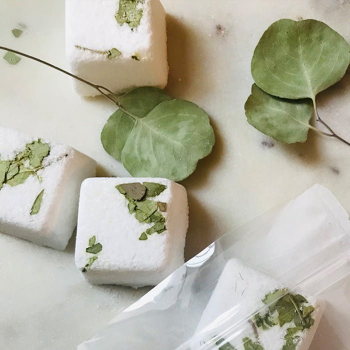 A closeup of the Eucalyptus and Peppermint Shower Steamers, lying on a cream surface next to itheir plastic packaging.