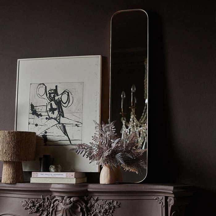 The Princeton Mirror, a thin rectangular mirror with curved corners leaning on a mantle behind a vase full of purple fern stems.