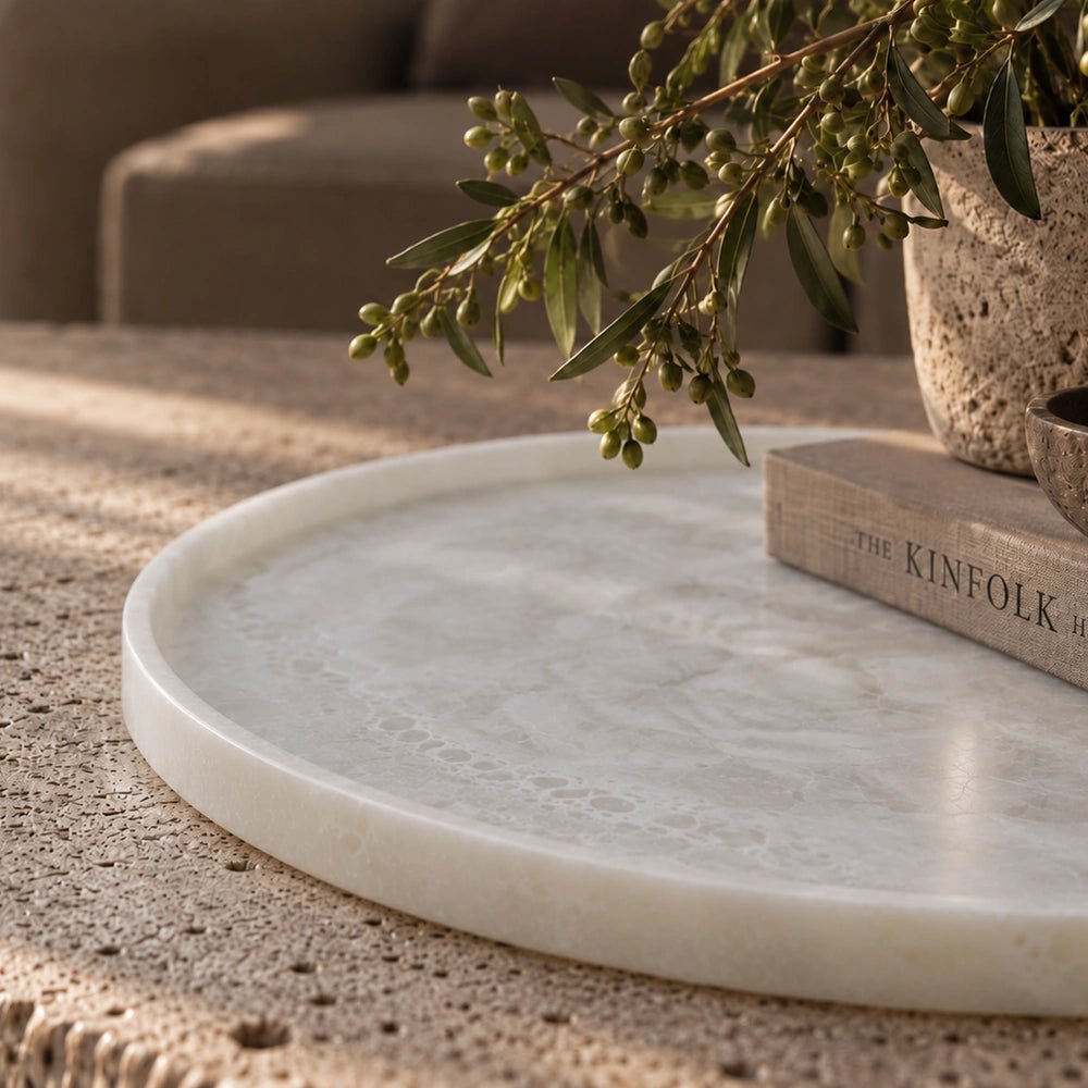 Salome Round Platter with smooth pale stone-effect surface and raised rim, shown close up on a textured tabletop beside books and leafy stems.