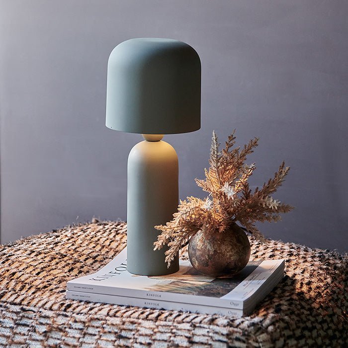 Sefton Table Lamp, a sleek matte sage metal table lamp with curved shade and smooth tapered base styled on a patterned pouf.