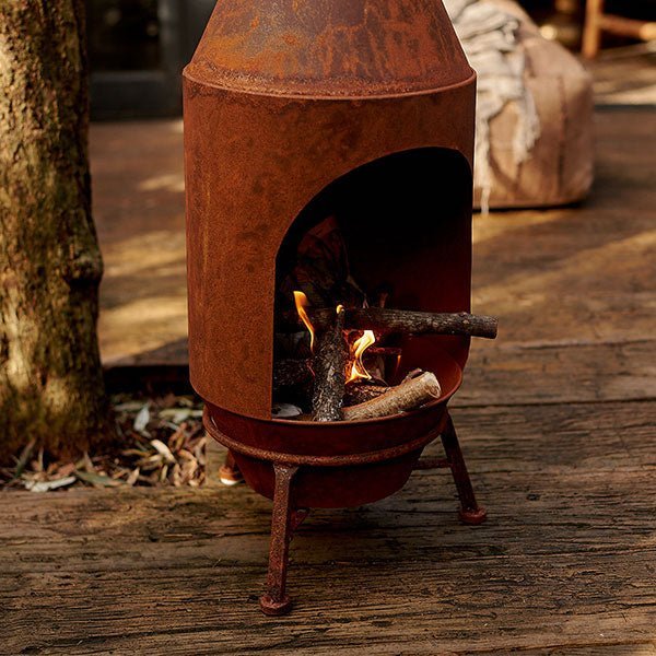 Sierra Chiminea Log Burner - Rust, an outdoor log burner in a classic shape and rust colour finish, on a shady wooden patio.