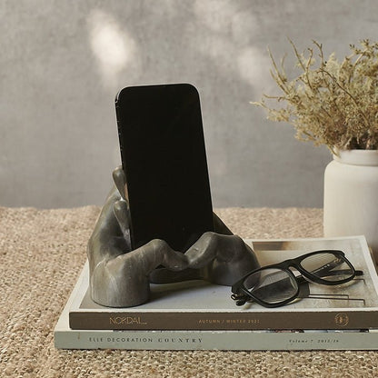 A phone placed in a grey phone holder shaped like two hands sat on a stack of books