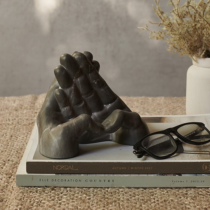 Grey phone holder in the shape of two hands sat on a stack of books next to a pair of glasses 