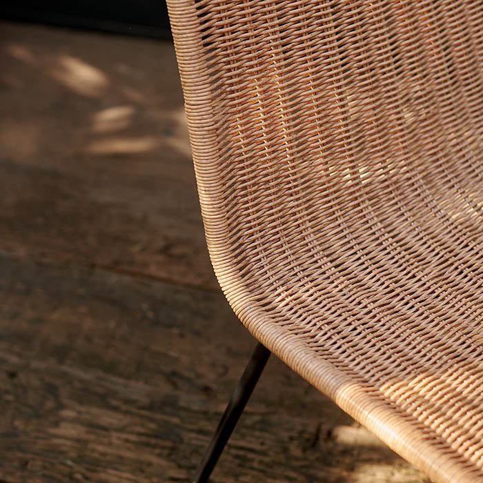 The Tatum Chair, a natural rattan woven chair in a contemporary style with slim black metal legs, on a sun dappled patio.