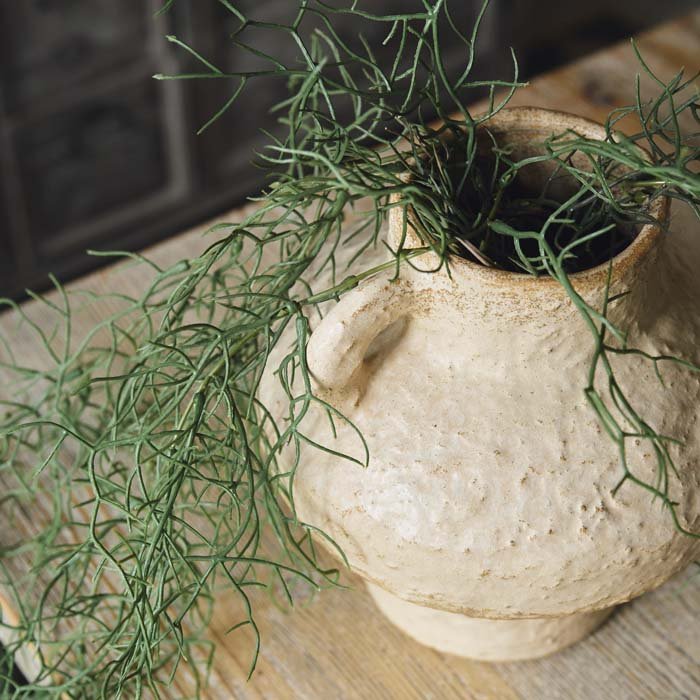 A couple stems of artificial spiky ferns trailing out of a beige ceramic vase.