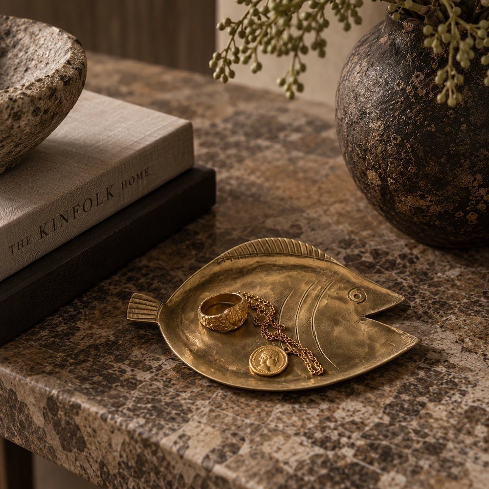 Torcino Brass Tray with irregular sculptural form and warm aged finish, styled on a textured table beside stacked books and dark decorative ceramics.