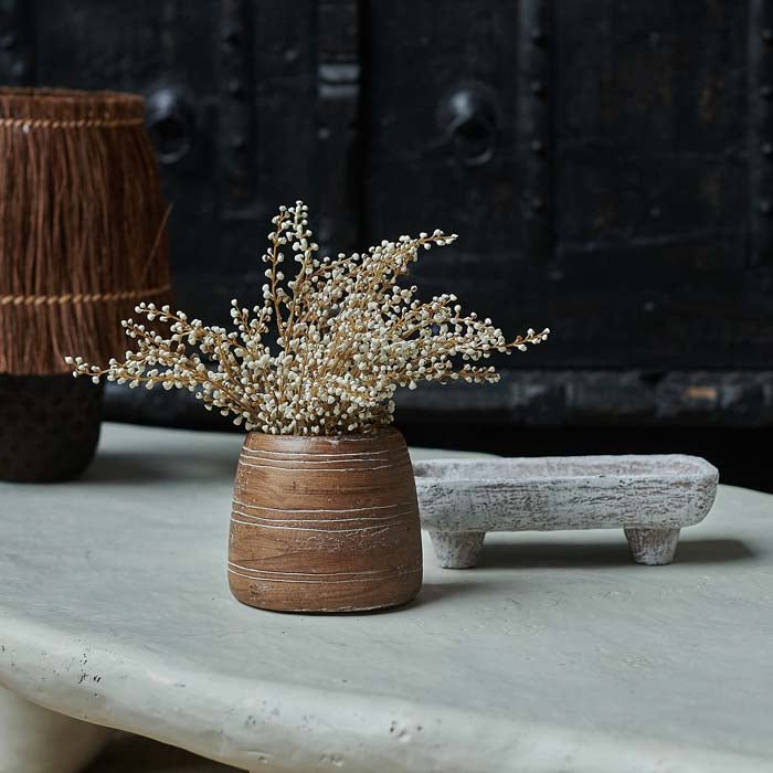 Round brown vase with ribbed markings filled with cream flower stems on a white coffee table  