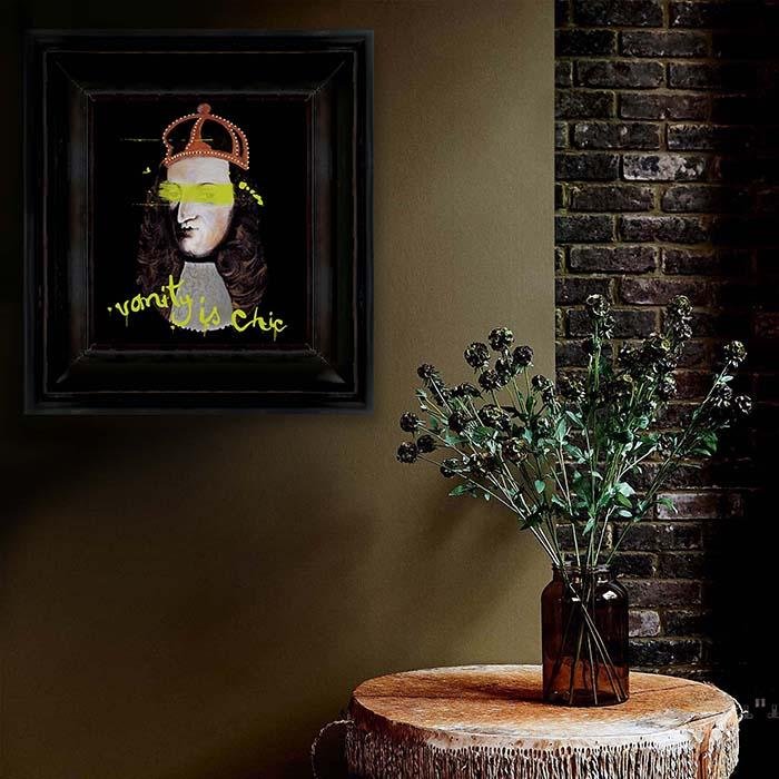 Face print with a red crown in a square thick frame hung on wall behind a vase