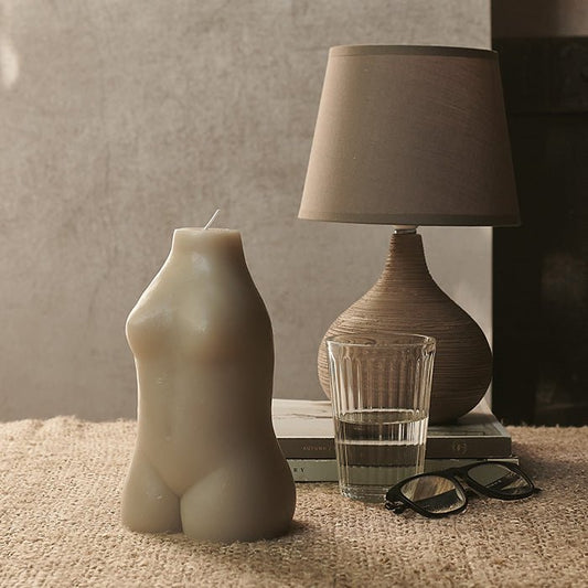 Female body shaped grey candle sat in front of a brown table lamp and a glass of water