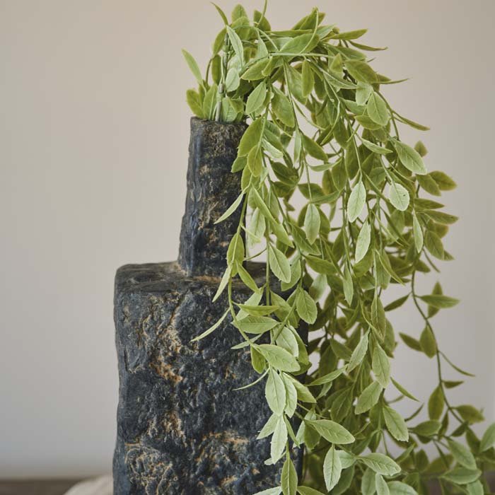 Detail image of a grey rustic vase holding a few trailing stems of fake weeping willow in a soft green colour.