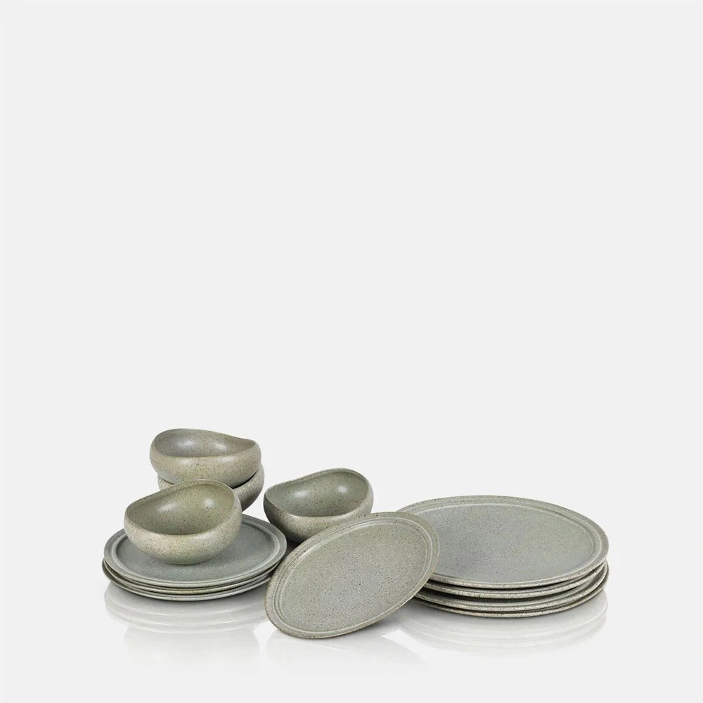 Alcott 12-Piece Dinner Set featuring pale green and beige speckled plates and rounded bowls, arranged in overlapping stacks with one plate leaning forward.
