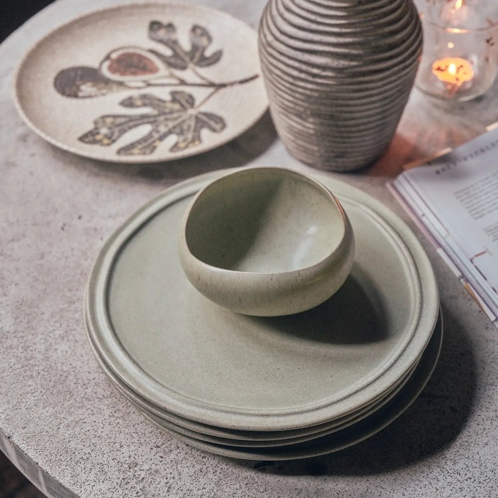 Alcott 12-Piece Dinner Set shown with a rounded green bowl resting on stacked plates, highlighting speckled surfaces and raised rims beside an open book.