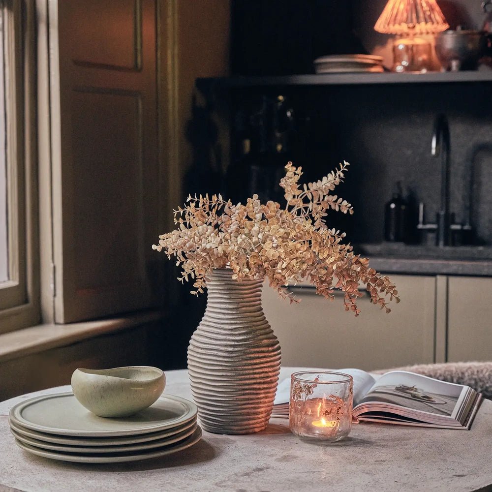 Alcott 12-Piece Dinner Set arranged with a green bowl atop stacked plates on a kitchen table, beside a botanical vase, glowing candle and open book.