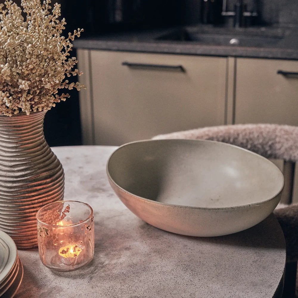 Alcott Serving Bowl with a wide rounded rim and gently curved grey interior, displayed beside a glowing candle and botanical vase on a kitchen table.