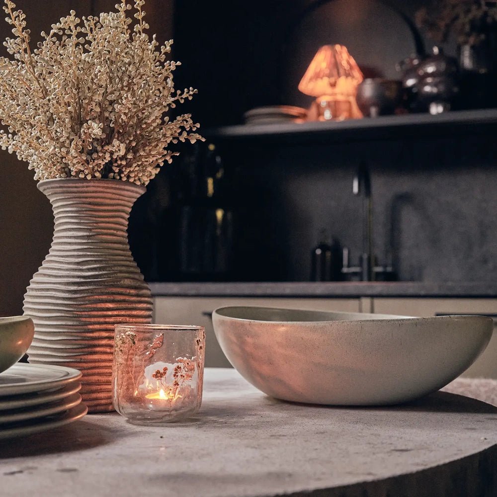 Alcott Serving Bowl with a broad rounded form and speckled grey surface, displayed on a kitchen table beside a glowing candle and botanical vase.