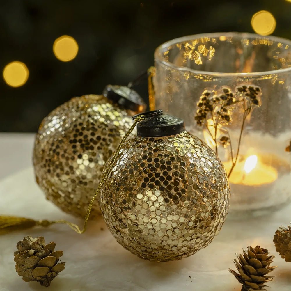 Alisa gold baubles with rounded shapes and reflective patterned surfaces rest beside a glowing candleholder and pine cones on a softly lit festive table.