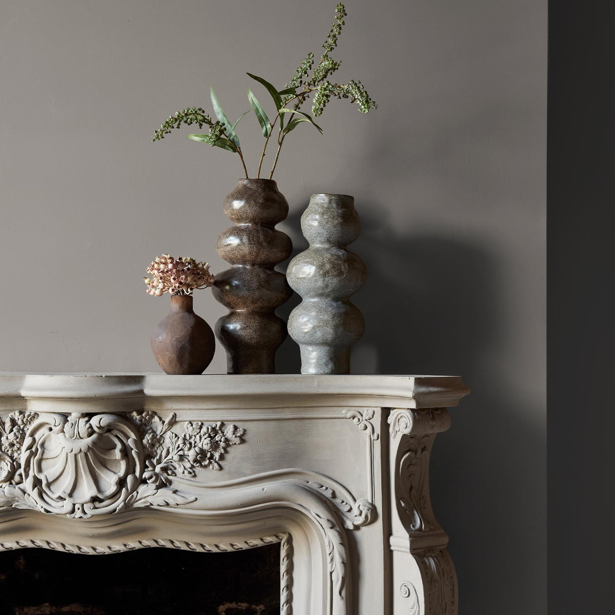 Three ceramic vases sitting on an intricate fireplace mantle in front of a wall in a light greige paint colour with beige undertones.