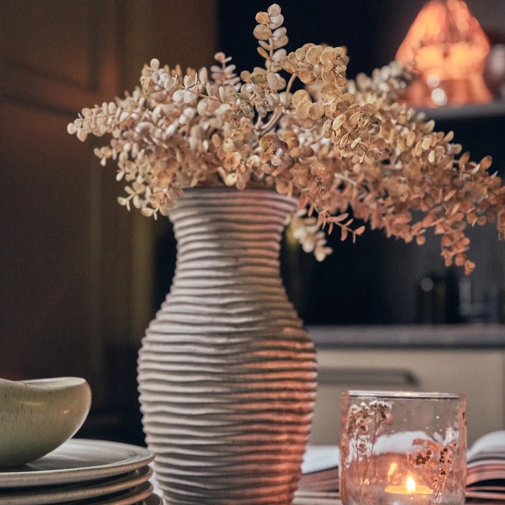 Ardley Stoneware Vase with a tall ribbed profile and flared neck, holding peach beige foliage on a dining table beside a candle and stacked plates.
