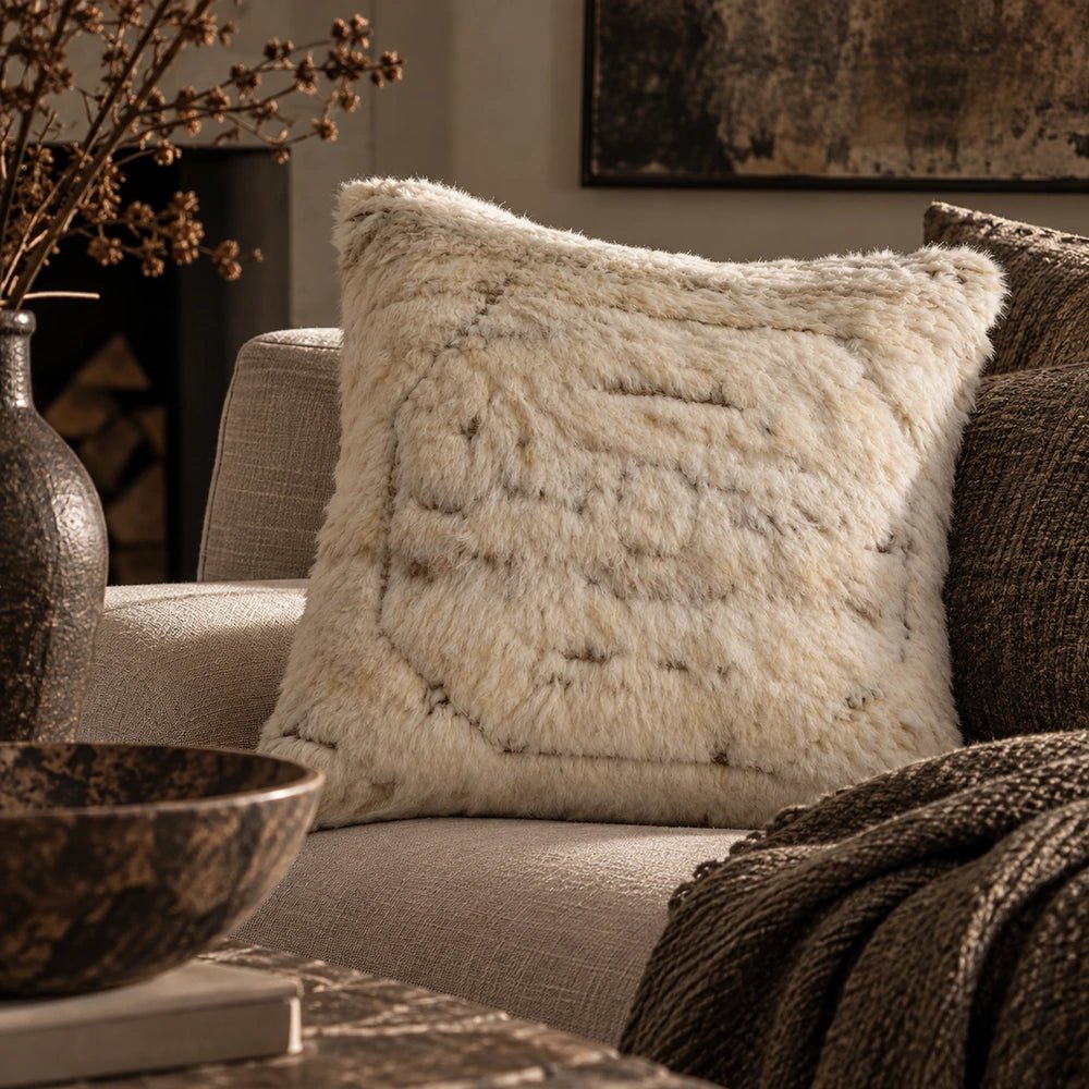 Astoria Wool Cushion with shaggy neutral texture styled on a dark sofa, layered with patterned cushions beneath oversized artwork in a contemporary room.
