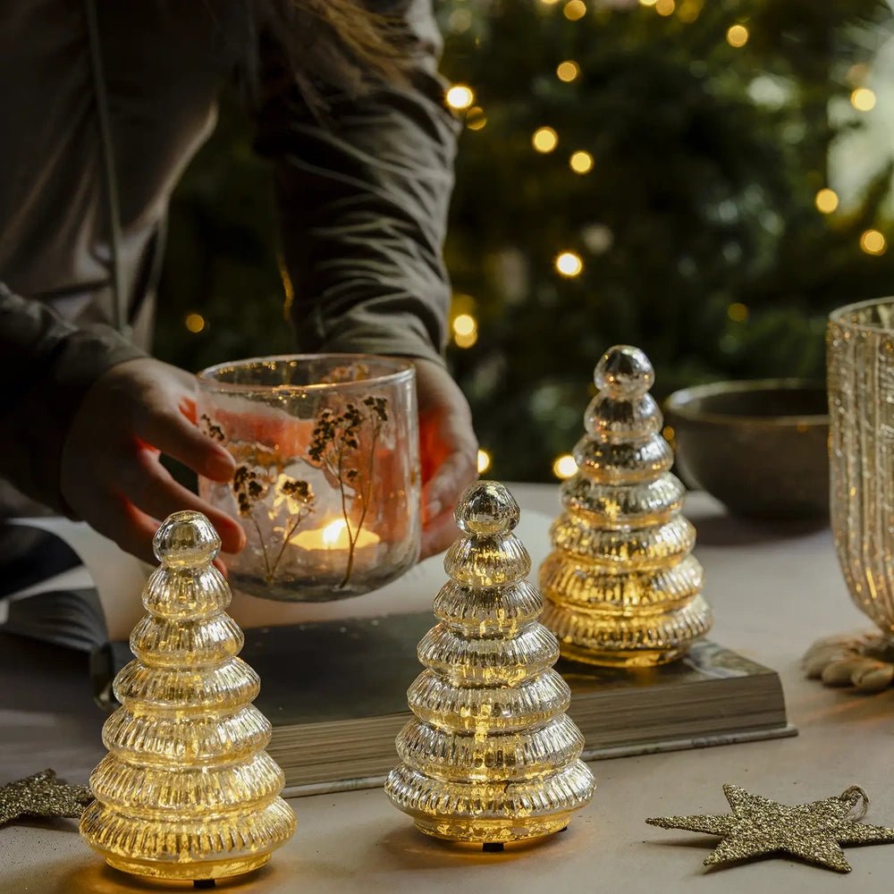 Aurora silver tree portable lamps cast a warm glow across a festive table while a person places a patterned candleholder beside their ribbed silhouettes.