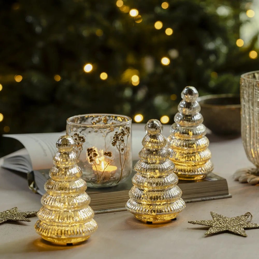 Aurora silver tree portable lamps in three sizes glow warmly through ribbed tiered surfaces, arranged on a festive table beside candles and star decorations.