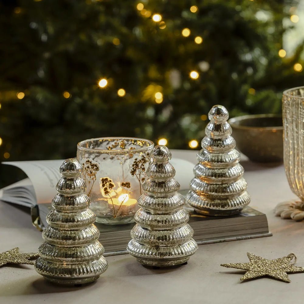 Aurora silver tree portable lamps in three sizes display reflective ribbed surfaces, arranged unlit beside a patterned candleholder and star decorations on a table.