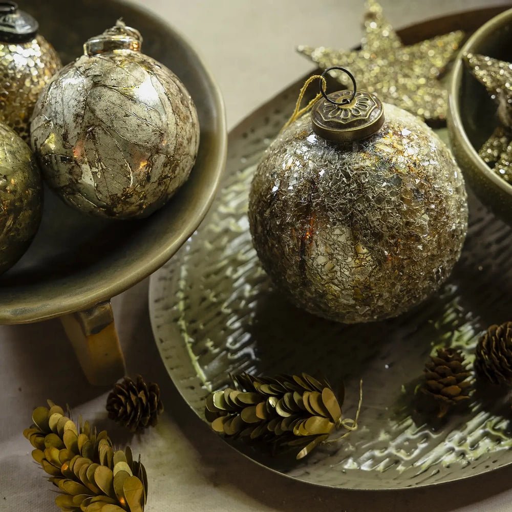 Beau copper flake bauble with a round mottled metallic surface and hanging cap rests in a decorative bowl beside pine cones and another ornament.