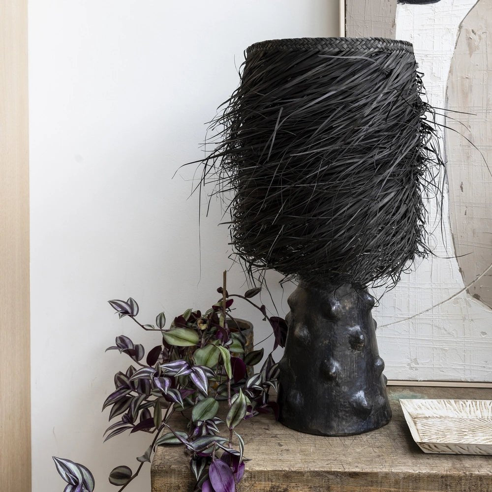 The Black Shaggy Palm Lamp has a tall fringed shade and textured sculptural base, displayed beside a trailing purple and green plant on a wooden console.