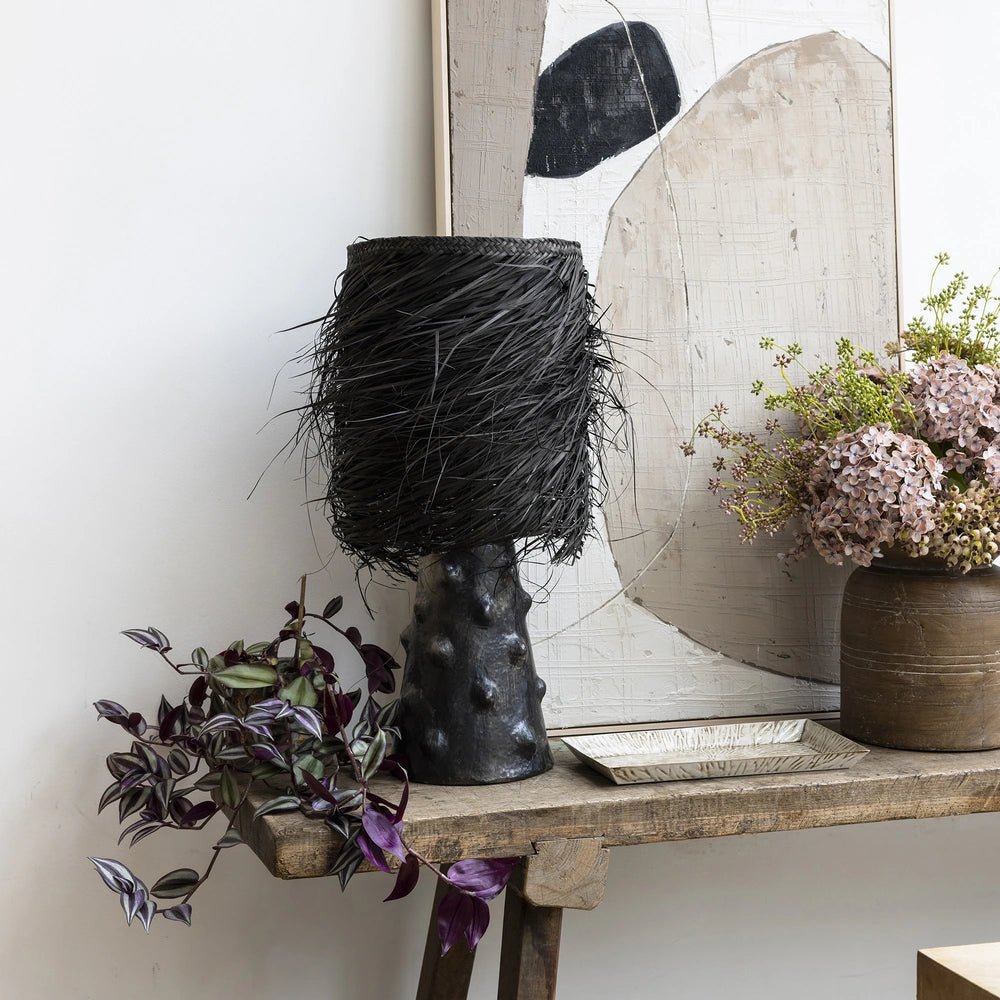 The Black Shaggy Palm Lamp stands on a rustic console with its woven fringed shade, dark textured base, trailing plant and pink floral arrangement.
