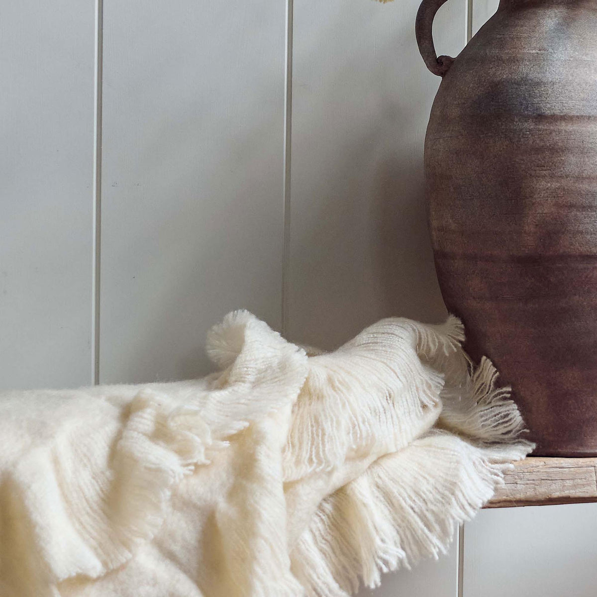 The Borough Beige Throw, a soft cream coloured throw with fringed ends on a low wooden bench next to a terrracotta vase.