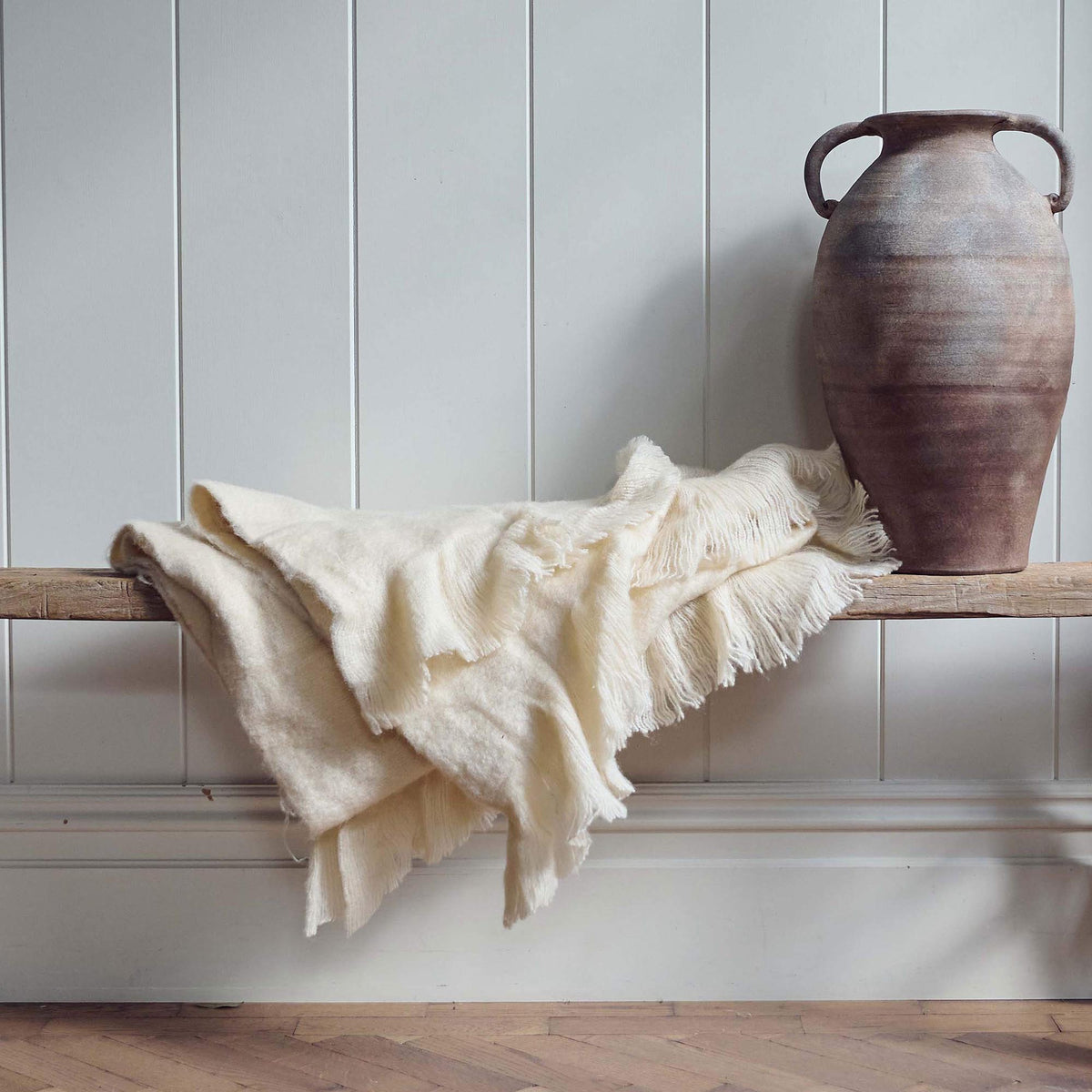 The Borough Beige Throw, a soft cream coloured throw with fringed ends on a low wooden bench next to a terrracotta vase.