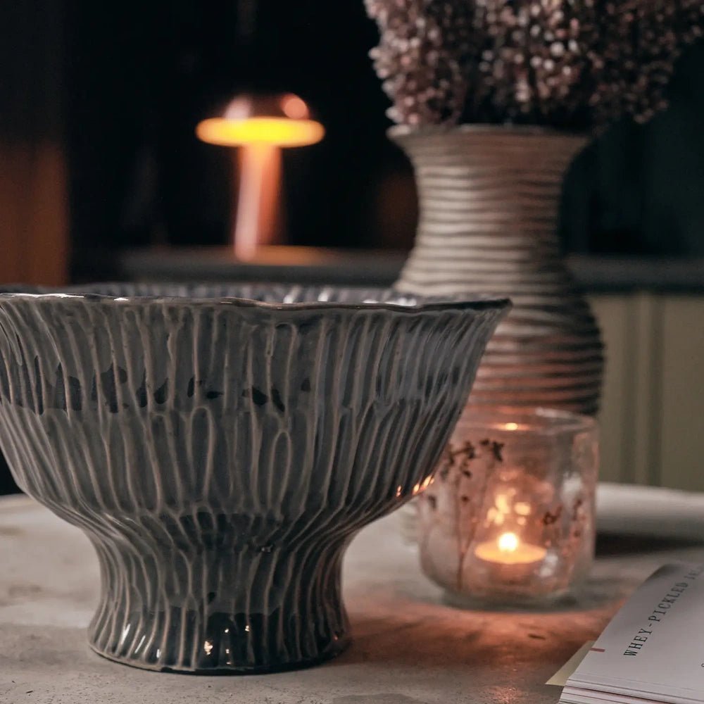 Bowham Ceramic Decorative Bowl with dark ribbed glazing and a raised foot, displayed on a tabletop beside a glass candle and a tall vase.