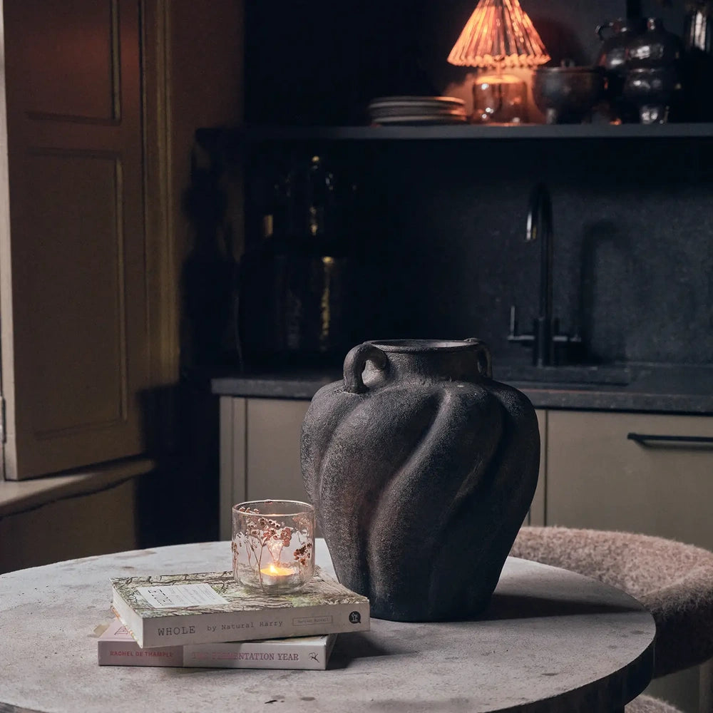 Braddock Magnesium Vase with a dark sculptural body and curved handles, styled with books and a candle on a round table in a moody interior.