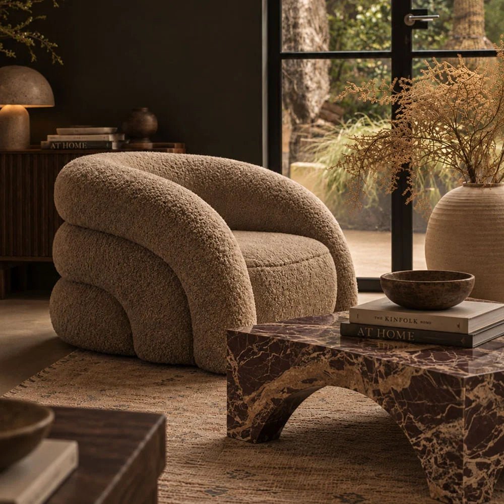 Braddow Marble Coffee Table with rich brown veining and curved arched legs, styled with books beside a cream armchair in a softly lit living room.