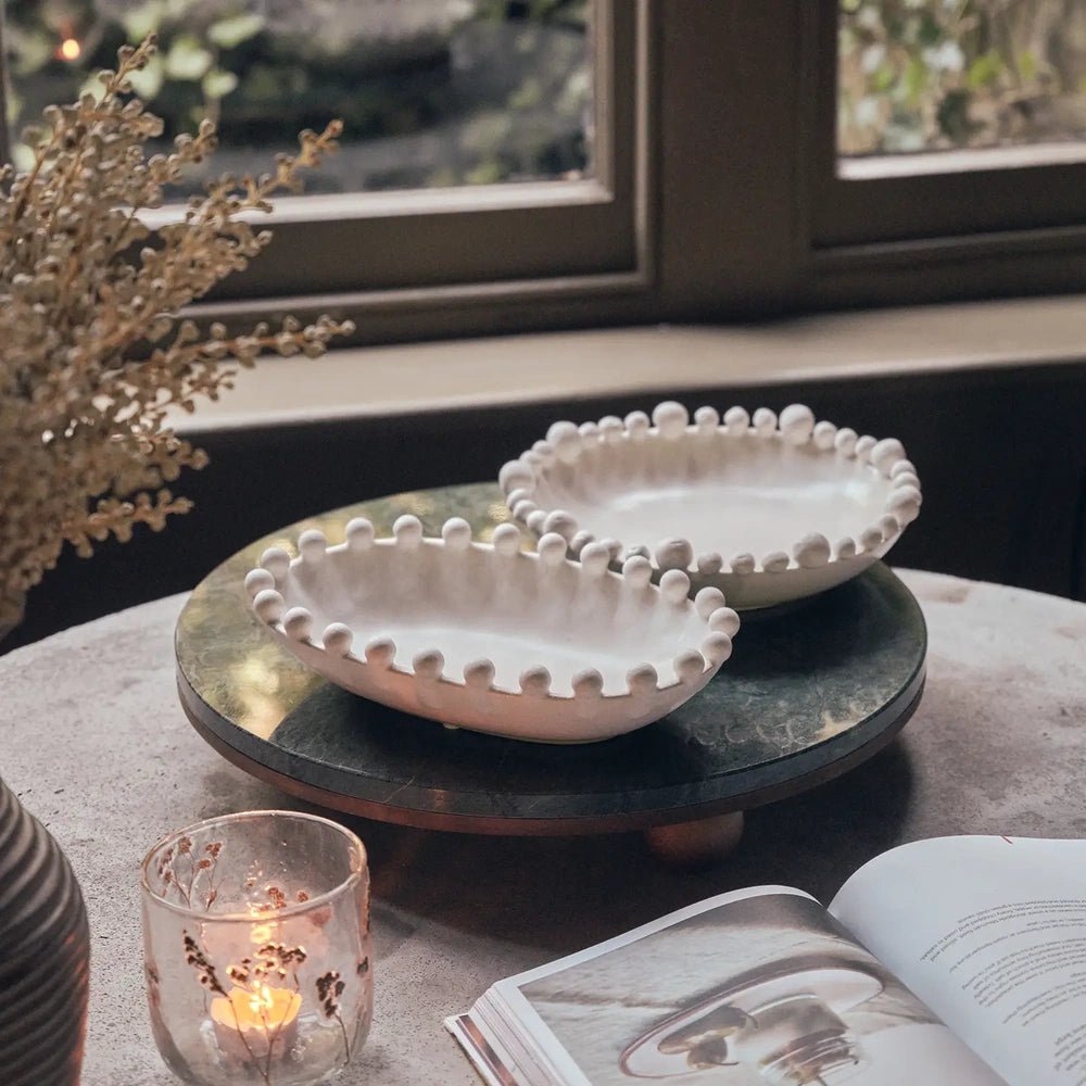 Brewster Bobble Bowl in a soft off-white finish with a rounded beaded rim, displayed with a matching bowl on a dark tray beside a window.