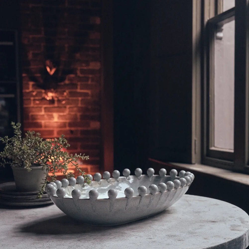Brigham Bobble Decorative Bowl in glossy grey with a rounded beaded rim, placed on a pale table beside a window and glowing brick fireplace.