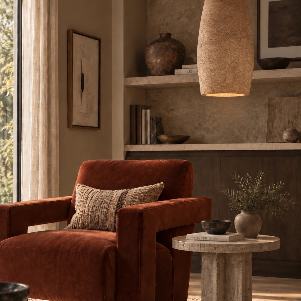 Cambria Paper Mache Pendant Light with tall textured shade, glowing above a warm neutral living room with rust armchair, stone table and layered shelving.
