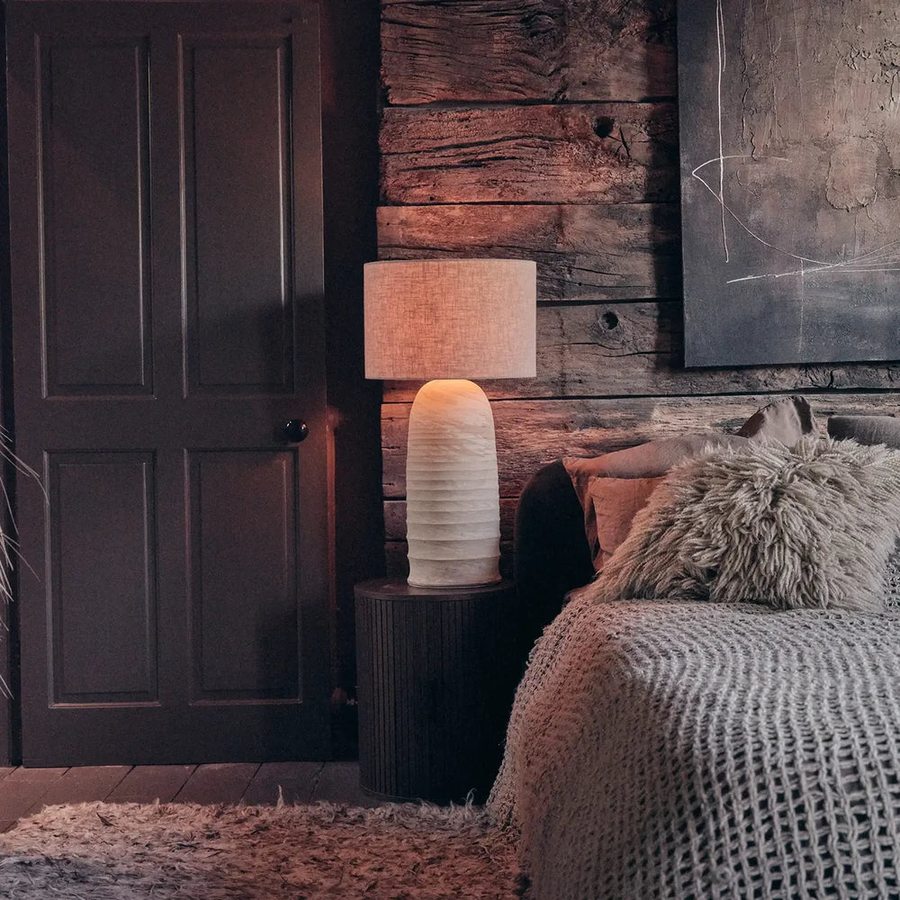 Campos Ceramic Table Lamp with a pale textured base and warm fabric shade, illuminating a dark wooden bedside table beside layered bedding and timber wall panels.
