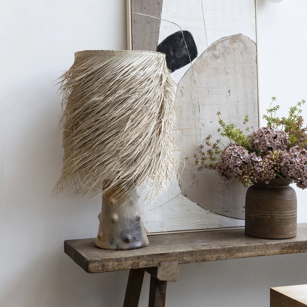 The Caramel Shaggy Palm Lamp has a pale fringed woven shade and sculptural textured base, displayed on a rustic wooden console beside pink flowers.