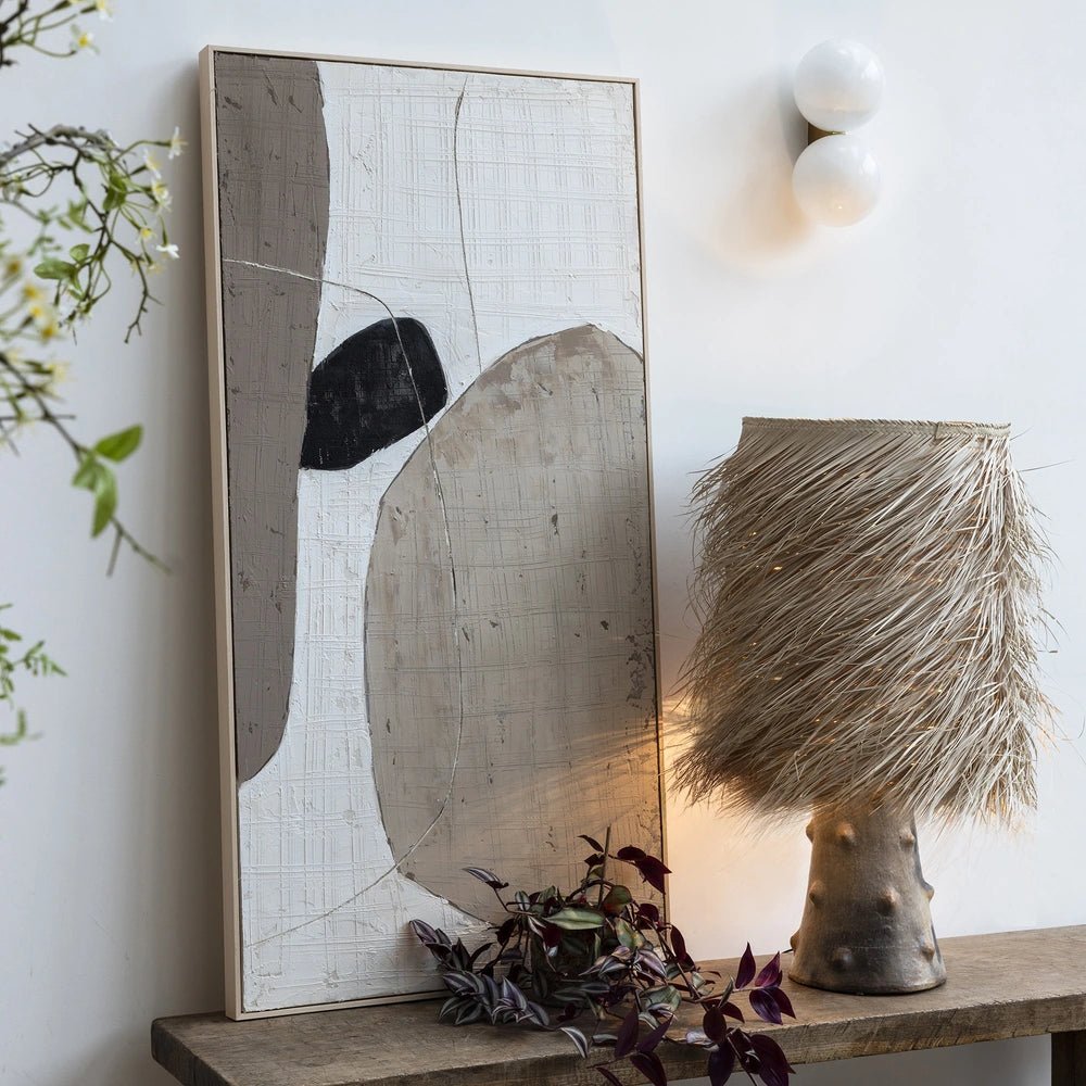 The Caramel Shaggy Palm Lamp glows through its fringed woven shade, with a textured sculptural base beside abstract artwork and trailing foliage on a console.