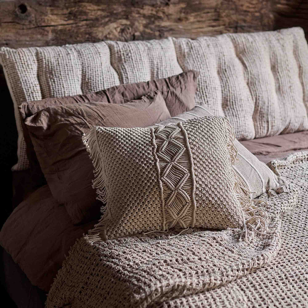 Cassidy Cushion, a square cushion with cream coloured macrame front and long fringe detail, next to a padded headboard.