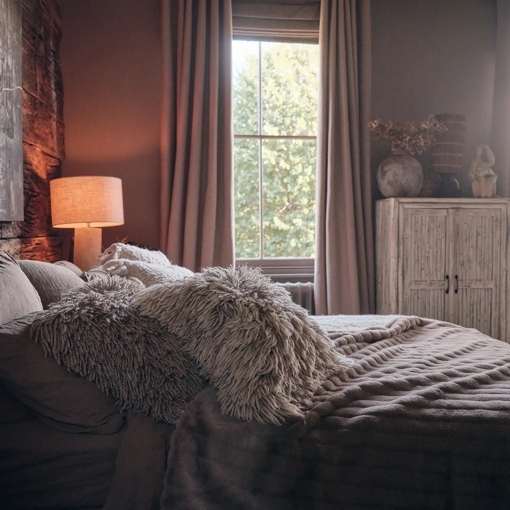 Chandler faux fur throw in soft taupe with plush ribbed texture, covering a bed beside a tall window, neutral curtains and a warm bedside lamp.