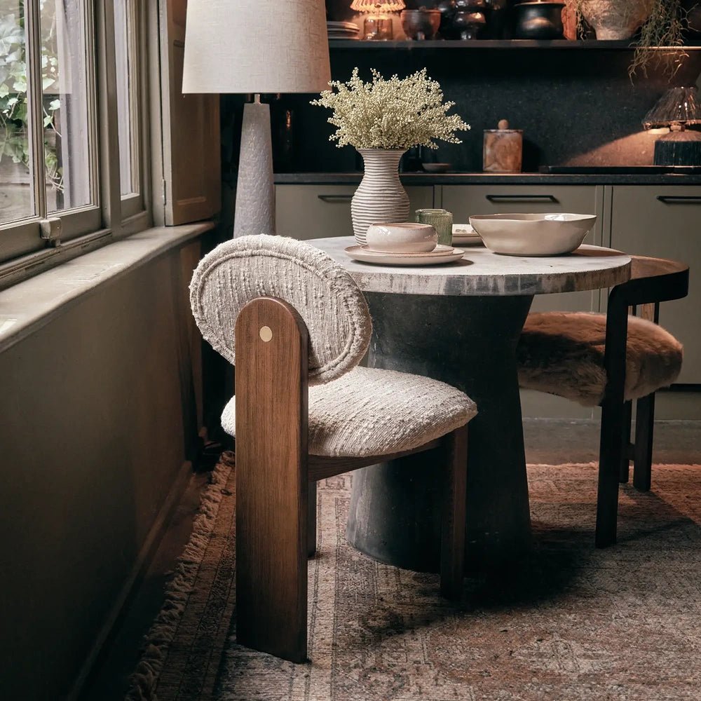 Channing boucle dining chair with a round cream backrest and wooden frame, positioned beside a circular dining table with ceramics, flowers and a floor lamp.