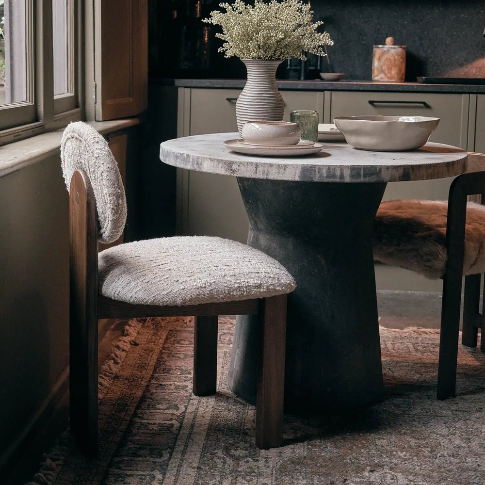 Channing boucle dining chair viewed from the side, showing cream textured upholstery and a dark wooden frame beside a round table near a window.
