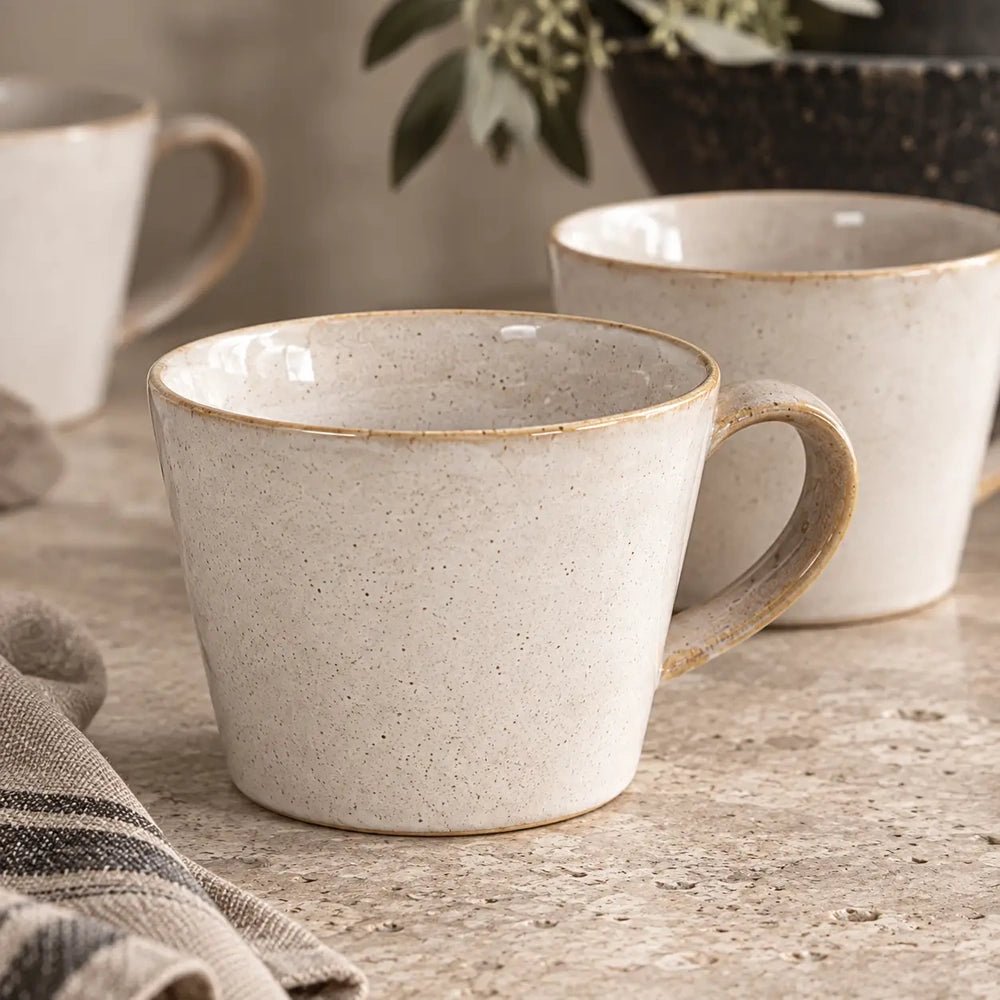 Coleman ceramic mug in warm off-white with a rounded handle and subtly speckled surface, arranged beside matching mugs and neutral linen on a stone counter.